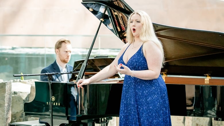 A woman in a blue dress stands in front of a piano singing.