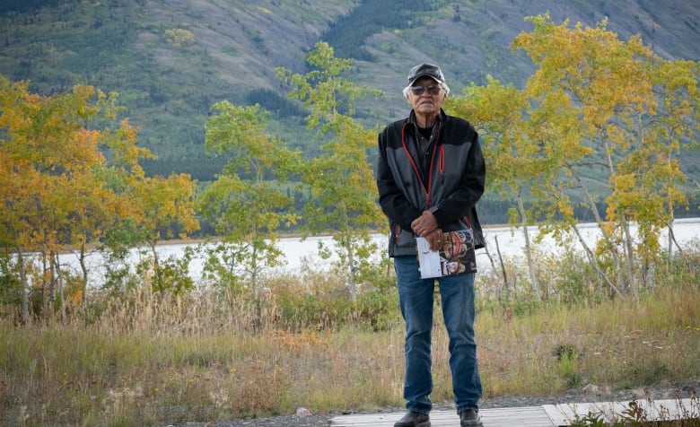 A man in a baseball cap and a dark jacket stands in front of a lake with a mountain in the background. 