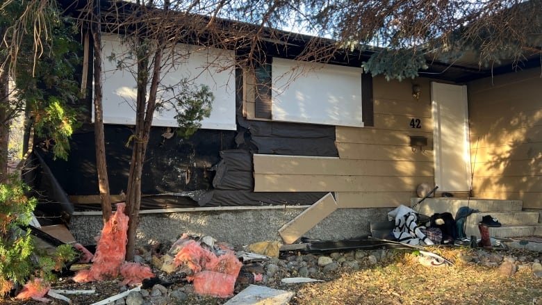 Pink insulation and other debris sits on the lawn in front of a house with damaged siding and a boarded-up door.