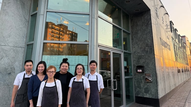 Six staff, mostly dressed in white shirts and black aprons, stand outside a restaurant.