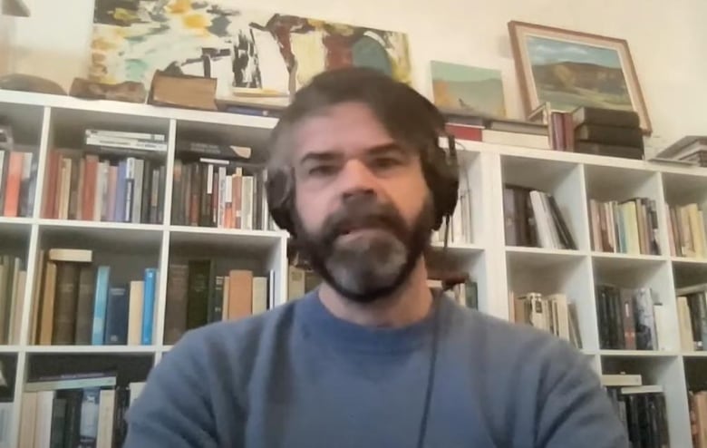 A man with a beard wearing a blue sweater and headphones sits in front of a bookcase.