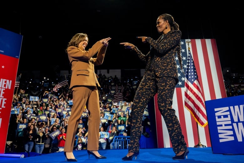 Two women in suits point at each other on an election campaign stage with an audience in the background.