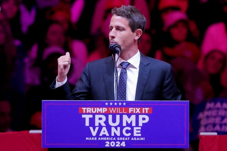 A dark haired man with mustache and beard is shown speaking at a podium that says Trump-Vance on it.