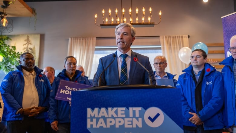 Man in blue suit stands at blue podium with several people standing at his side