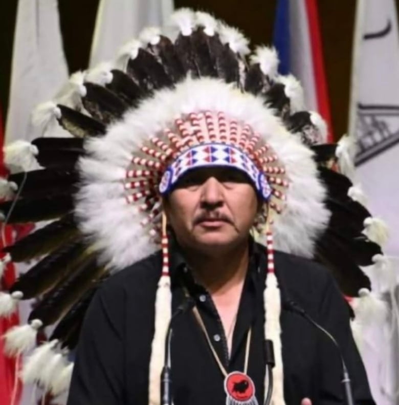 A mean in a headdress speaks at a podium.