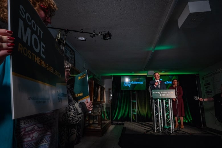 A man and a woman stand behind a podium in the distance. In the foreground, a person holds an election sign.