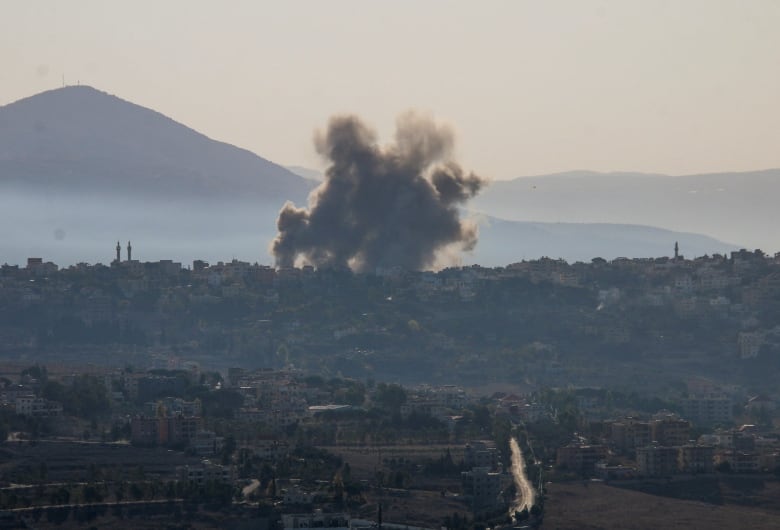 A large cloud of smoke is shown from a distance rising above a town located near a hill or mountain.