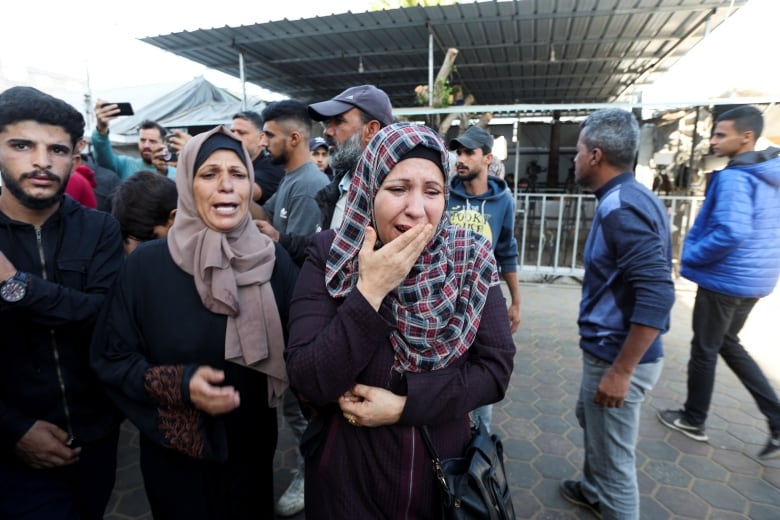 Two women wearing headcoverings are prominent in a photo, standing outside with pained expressions. Several others, mostly men, are seen in the background.