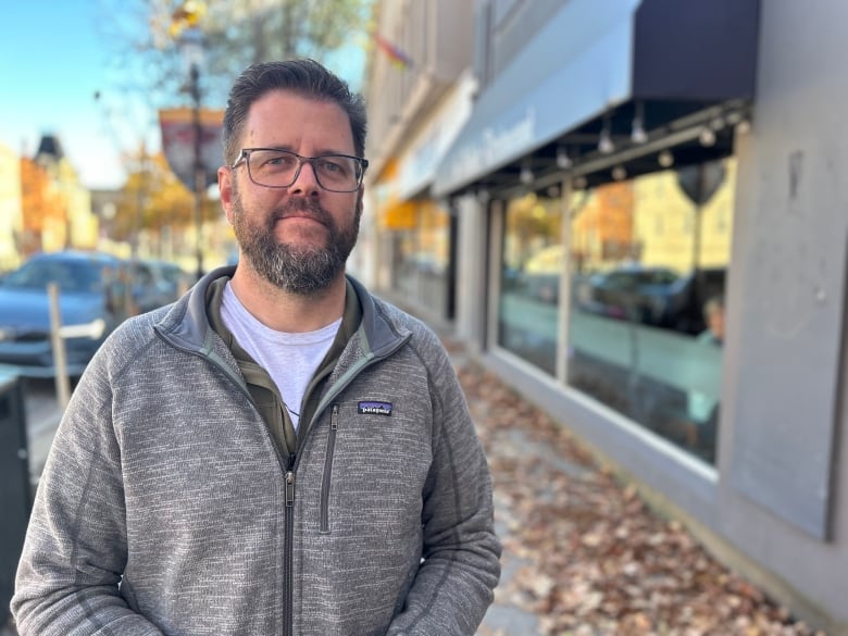 A man with brown hair, a beard and glasses smiles at the camera with fall leaves and a building in the background.