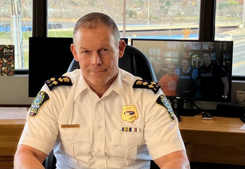 A clean shaven man with short hair, dressed in a police uniform, sits at a desk in front of a window. 