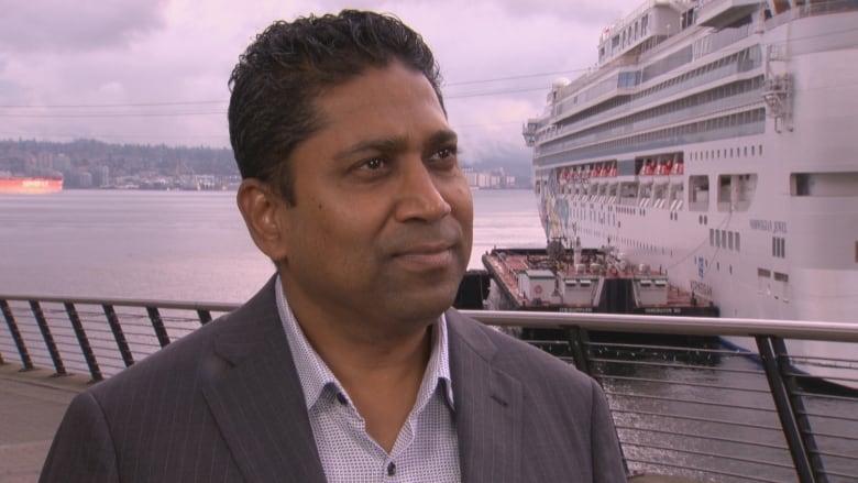 A young man in a suit and open-collared shirt speaks to a reporter with a docked cruise ship behind him in the harbour.