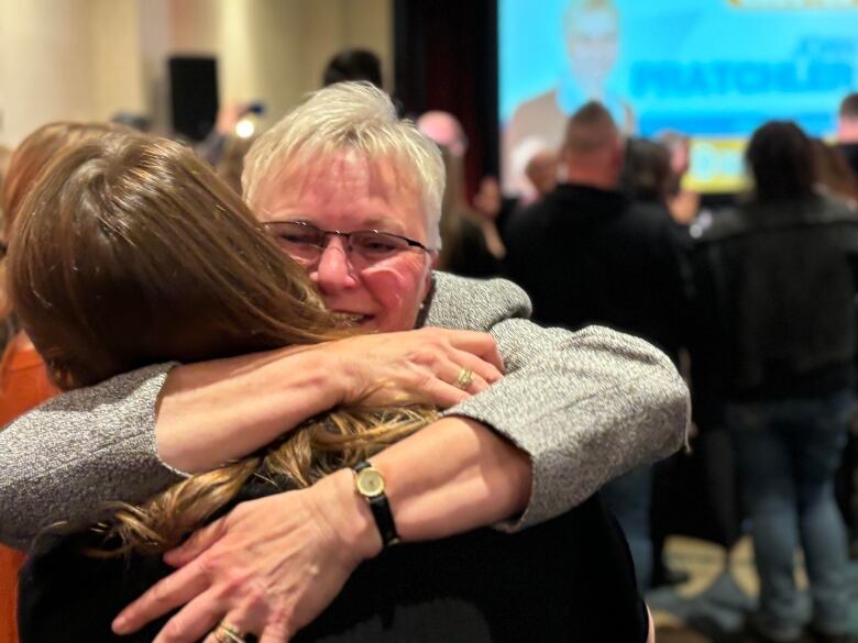 A woman in a grey suit jacket hugs a woman in black.