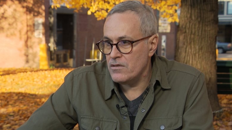 A man with round glasses sits at a table outside in the fall.