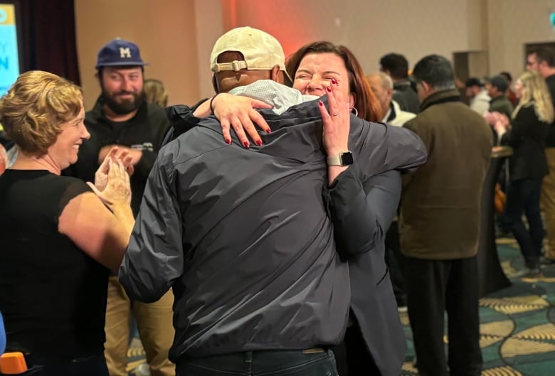 A woman in grey hugs a man in a hat. 