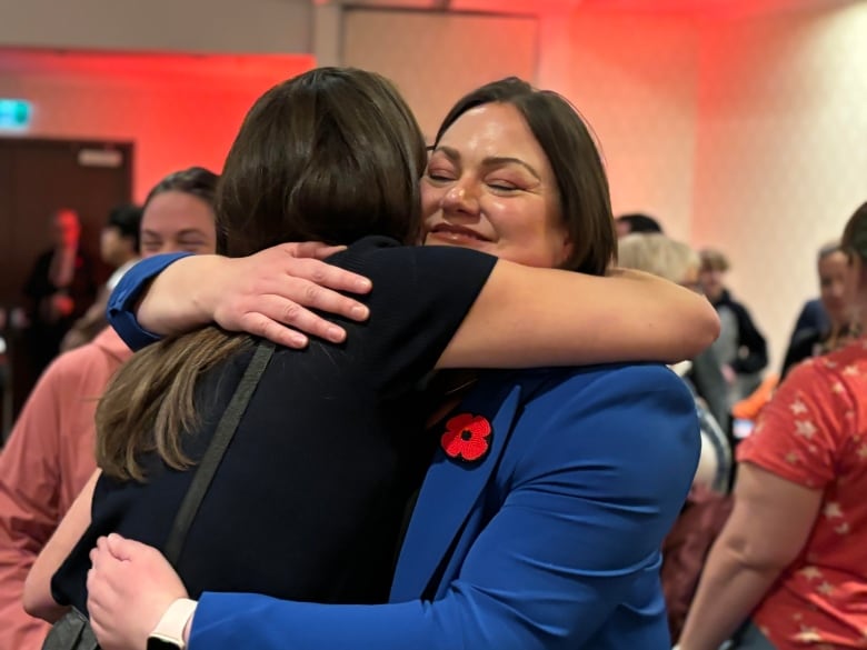 A woman in a blue suit with a red poppy hugs a woman in black. 