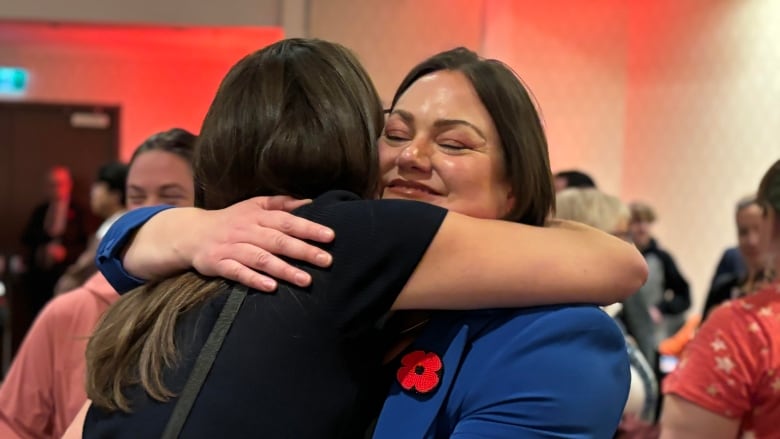 A woman in a blue suit with a red poppy hugs a woman in black. 