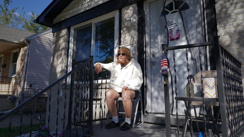 A woman in a white hoodie, black shorts and black sunglasses sits on a porch in front of a white house on a sunny day.