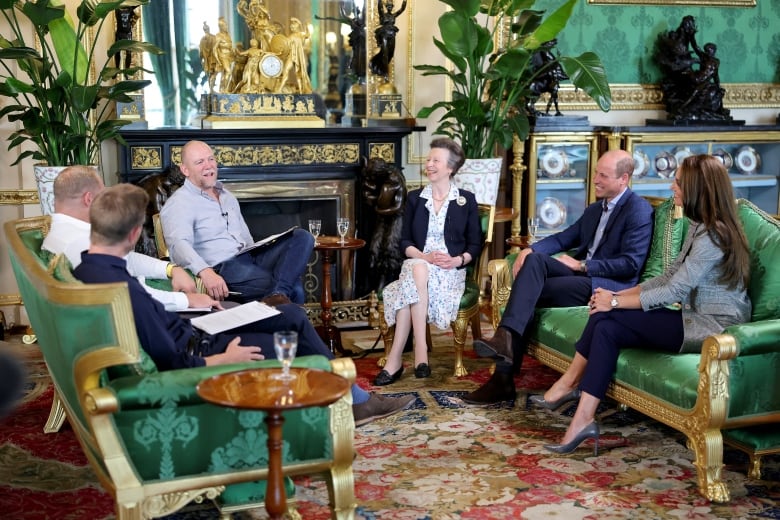 Six people seated on sofas and chairs in an elaborate living room talk with one another.