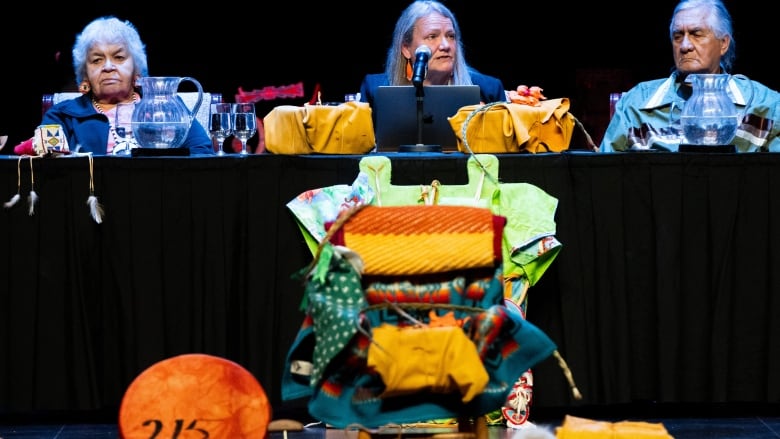 An empy chair draped with blankets sits in the foreground while people sit at a podium in the backgroun.