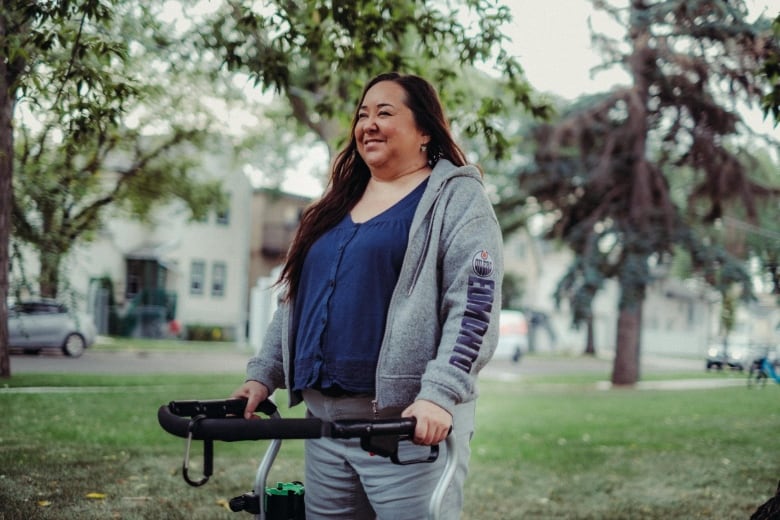 A woman smiles as she stands with a walker with trees behind her in an urban park.