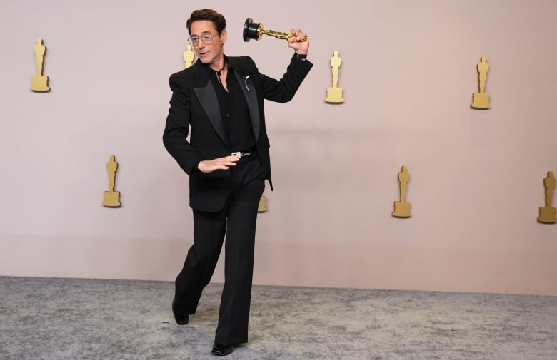 A man is shown on a velvet carpet in a suit. He has one leg kicked bent behind him in a jaunty fashion and is gesturing with his Oscar statue as though he is going to throw it or swing it like a bat or break into a dance. 