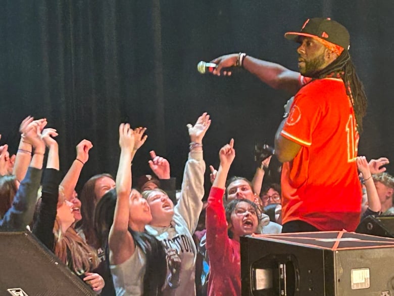 Crowd singing in a mic pointed towards them by a black man with black braids, wearing a hat and red jersey.