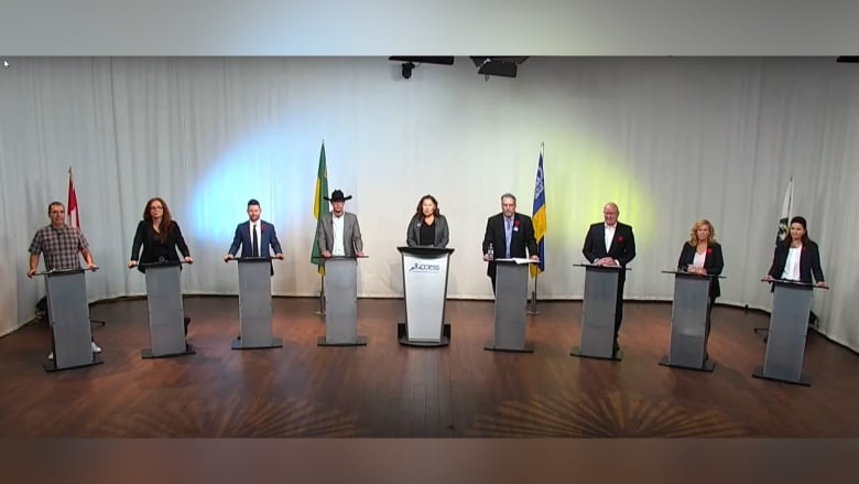 Lined up in a row are nine people standing behind podiums and in large room with wood floors. 