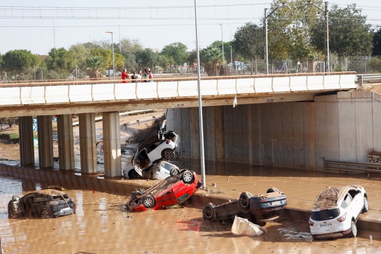 Two overturned cars and three other vehicles are shown in muddy water by a bridge.