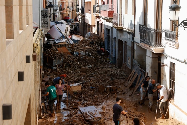 A narrow alleyway in a town is filled with people with brooms and mops, as well as debris, mud and damaged vehicles.