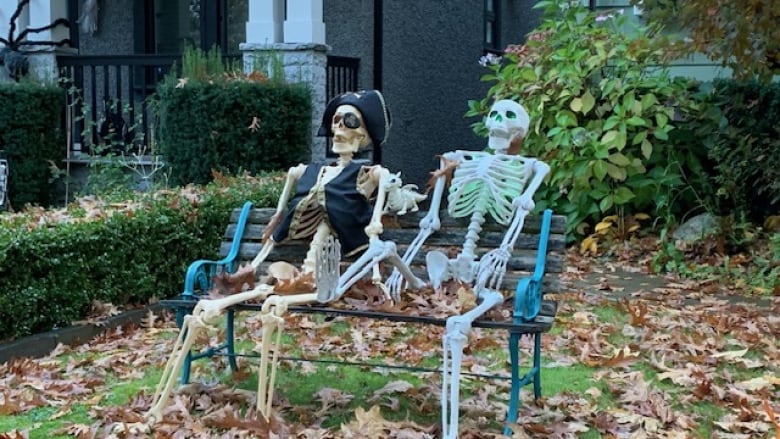 Two life-size skeleton decorations sit on a bench on a lawn.