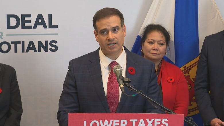 A man in a dark suit with a red tie speaks at a podium while a woman in a red blazer in front of a Nova Scotia flag stands behind him. 