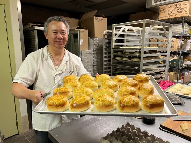 A man holds a tray of golden buns in a commercial kitchen.