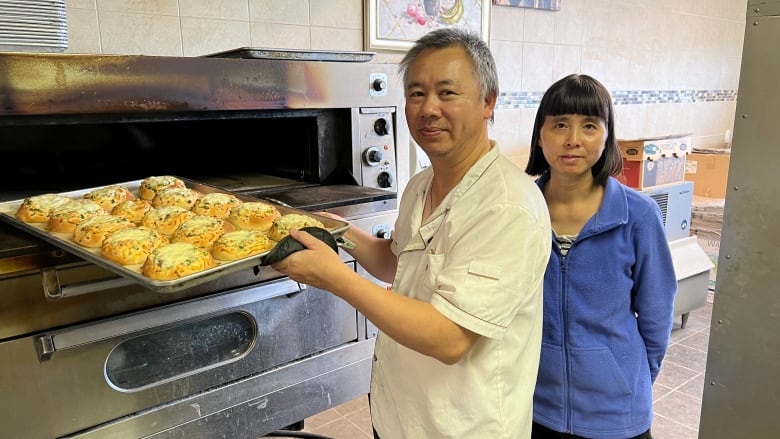 A man holds a tray of buns for baking in front of a large oven, while a woman with black hair stands beside him.