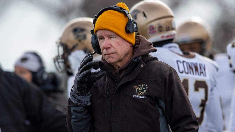 A football coach in a yellow tuque and headset watches from the sideline with players behind him.