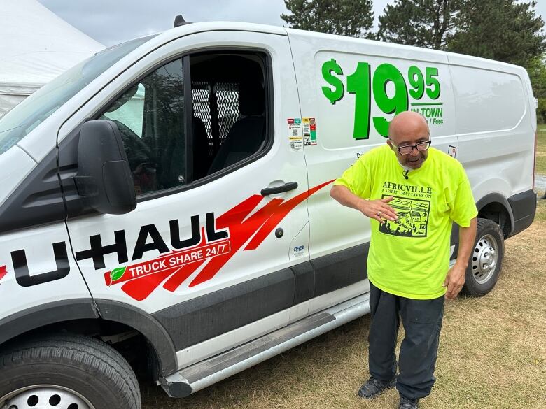 A man in a yellow Africville T-shirt is shown outside a white rented van in a park.