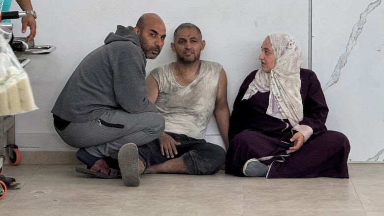 A wounded man sits on the ground in between another man and a woman.