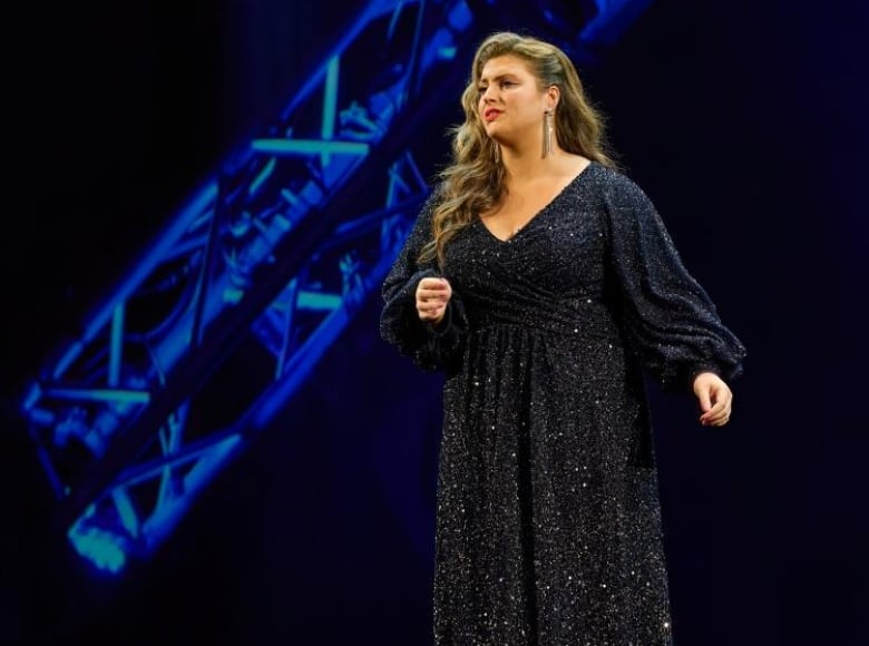 A woman with blond, curled hair, wearing a black sparkly gown, singing on a stage.