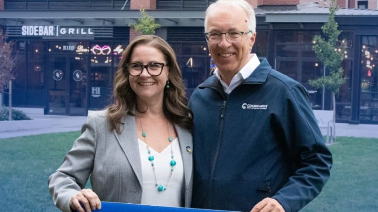 A woman with glasses and brown hair stands with a man with grey hair and glasses. They hold a  B.C. Conservative Party sign that reads Misty Van Popta.