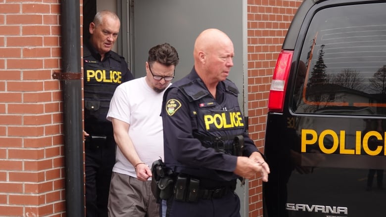 A man wearing glasses and a white T-shirt, as well as handcuffs, is escorted into an OPP van by two officers.