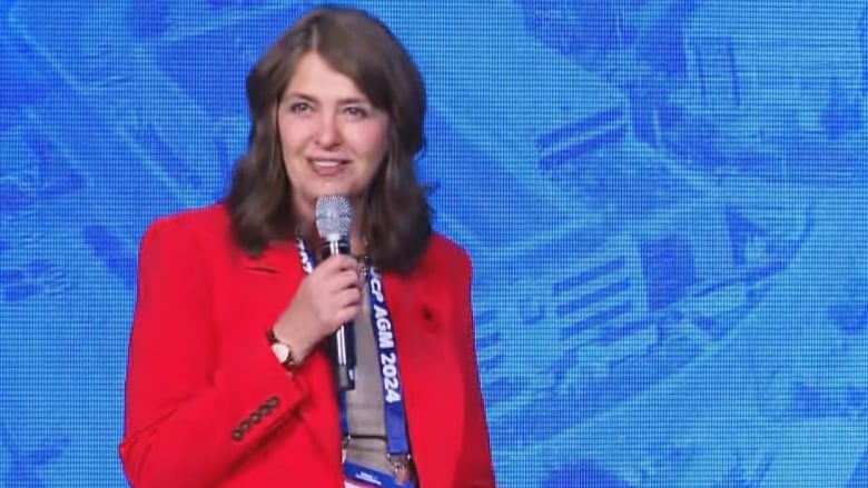 A woman in red speaks into a microphone in front of a blue screen. A man is scene on the right side.