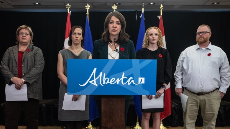 A brown haired woman stands at a podium as four people stand behind her in a room with Alberta and Canadian flags.