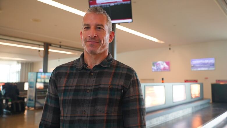 A man with short grey hair smiles as he stands in front of a baggage carousel.