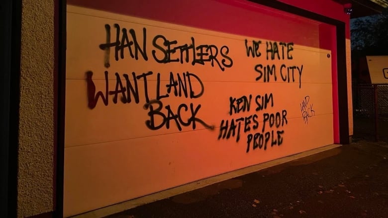 Scrawled text on a garage door reads 'Han Settlers Want Land Back' 'Ken Sim hates poor people' 'We hate Sim City' ' Land back'.