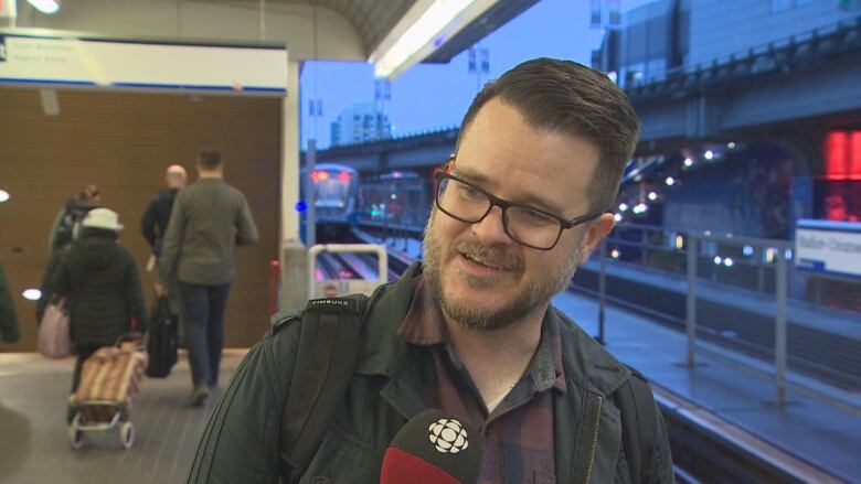 A man at a train station smiles, with others in the background.