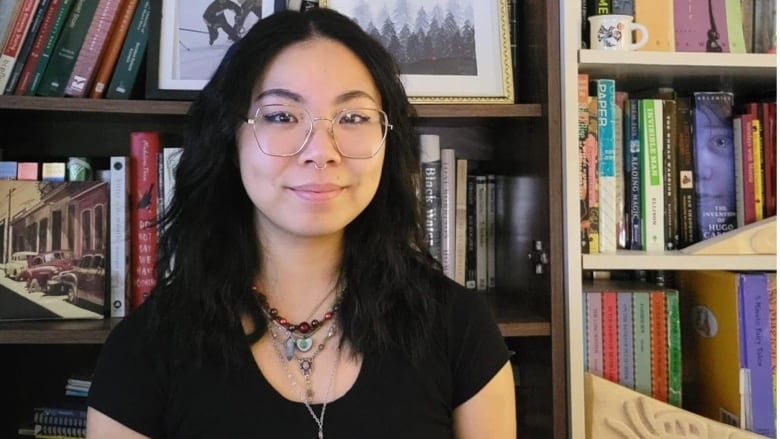 An Asian woman with glasses and long black hair wearing a black top and multiple overlaid necklaces sitting in front of a bookshelf