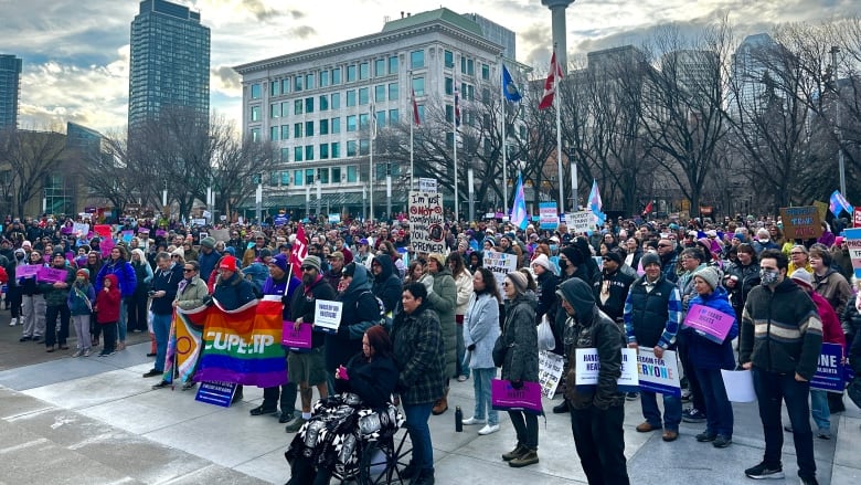 A large crowd of people outdoors bearing various signs and blue and pink transgender flags.