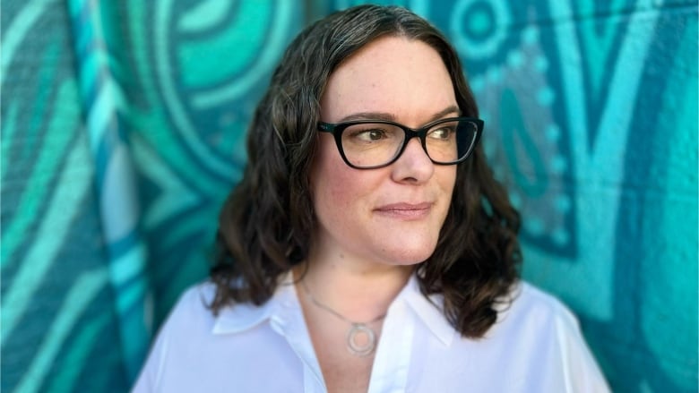 A woman wearing a white blouse and with shoulder length brown hair and dark rimmed glasses is standing in front of a teal wall while looking to the side