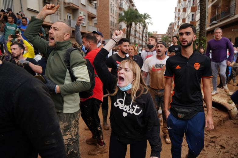 People stand in a flooded street and some of them are yelling and raising their fists in the air. 