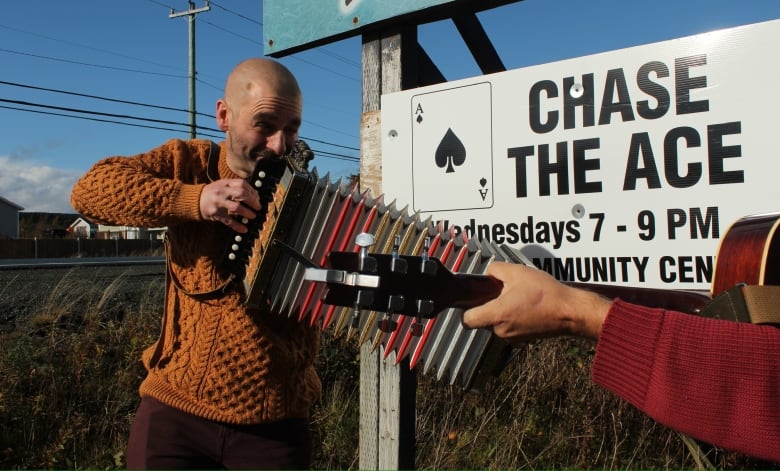 Man in orange knit seater playing an accordion. There is a sign that says Chase the Ace behind him.