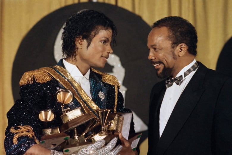 Michael Jackson holds several Grammy awards in his arms and smiles at another man, wearing a tuxedo.